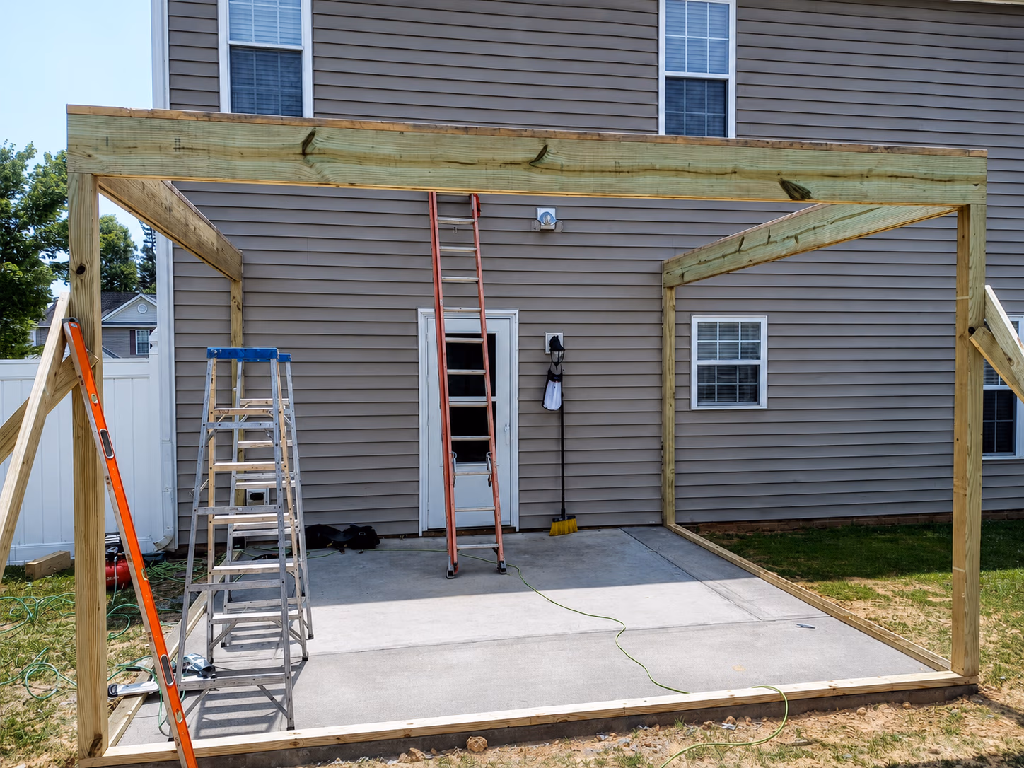 Wooden framework structure under construction on concrete patio outside brown house with ladders and tools.