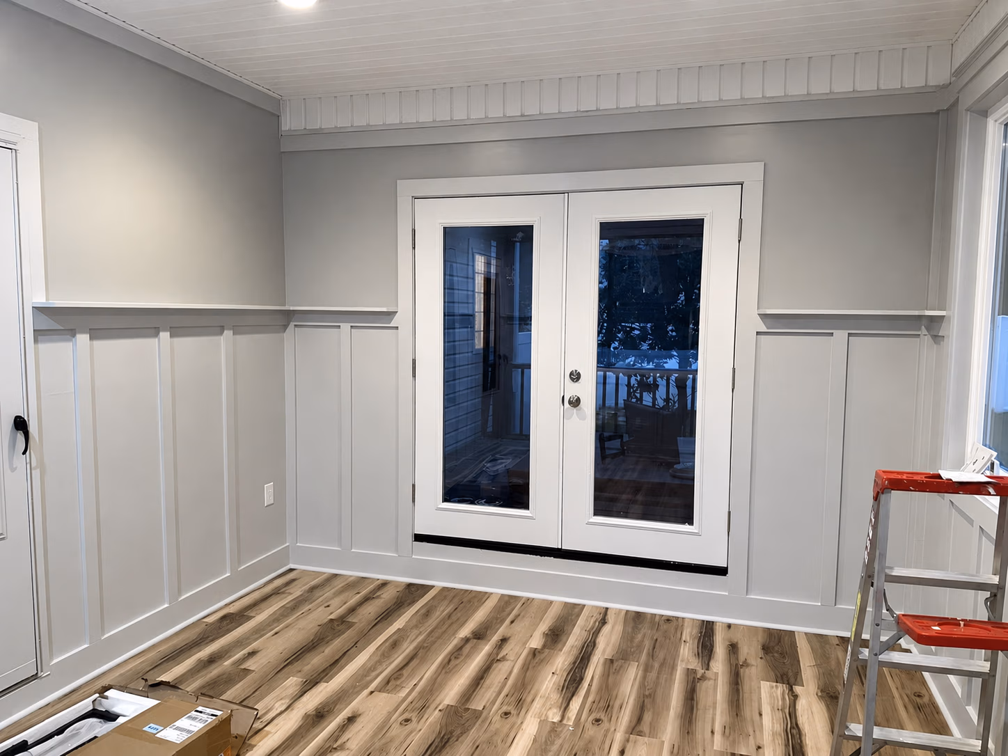 Empty room with light gray wainscoting, wood floor, white ceiling, French doors, and a ladder with a red tray.