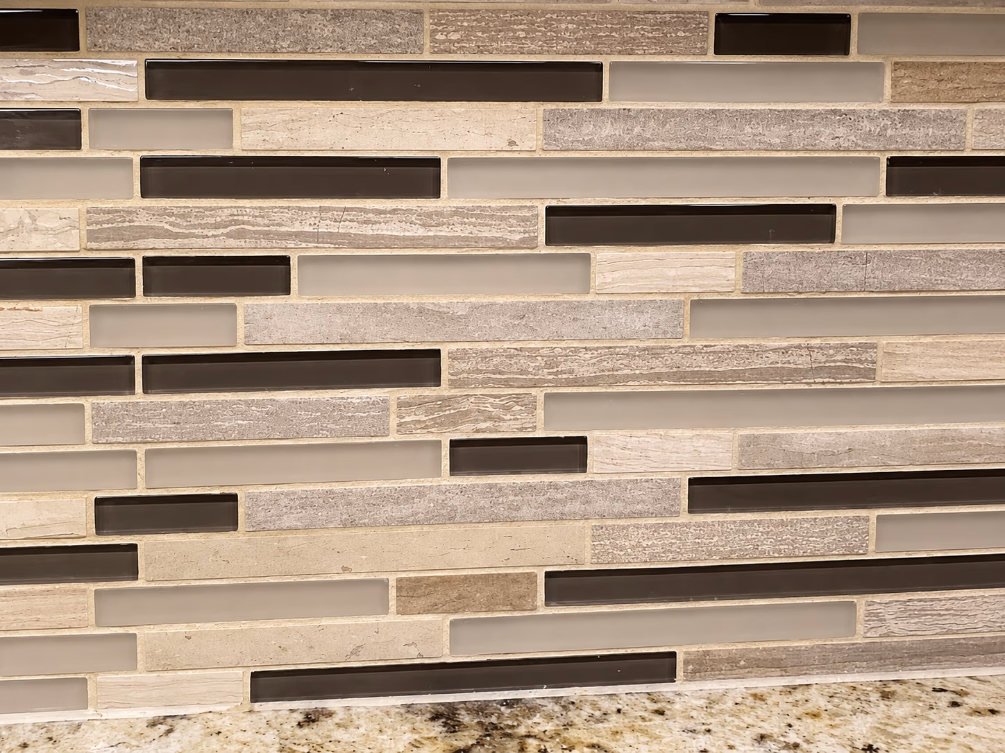 Kitchen backsplash with mixed beige and brown rectangular glass and stone tiles above granite countertop.