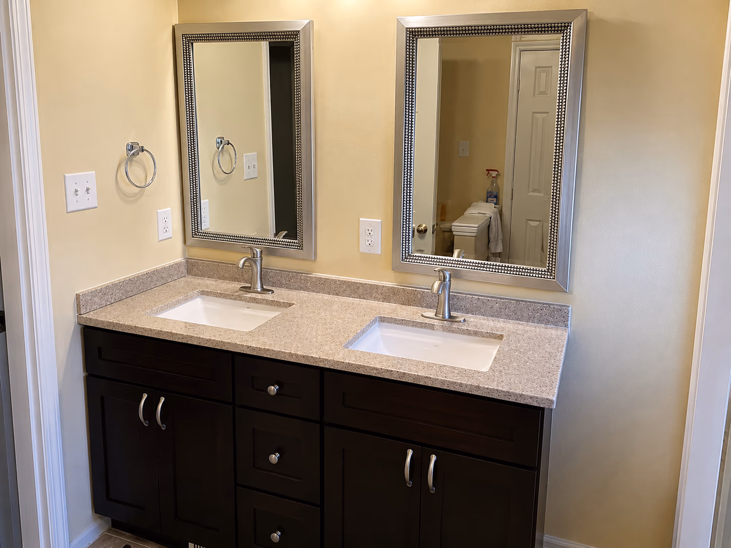 Bathroom vanity with two sinks, silver faucets, dark cabinets, and two framed mirrors on a beige wall.