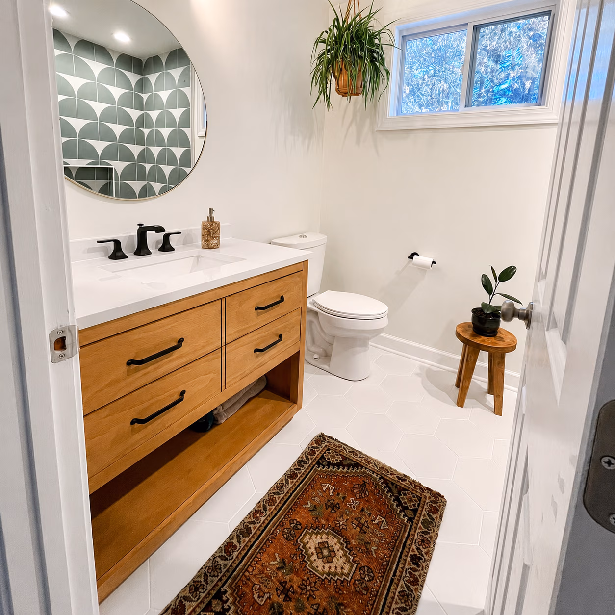 Modern bathroom with wooden vanity, round mirror, white toilet, patterned rug, and potted plants.
