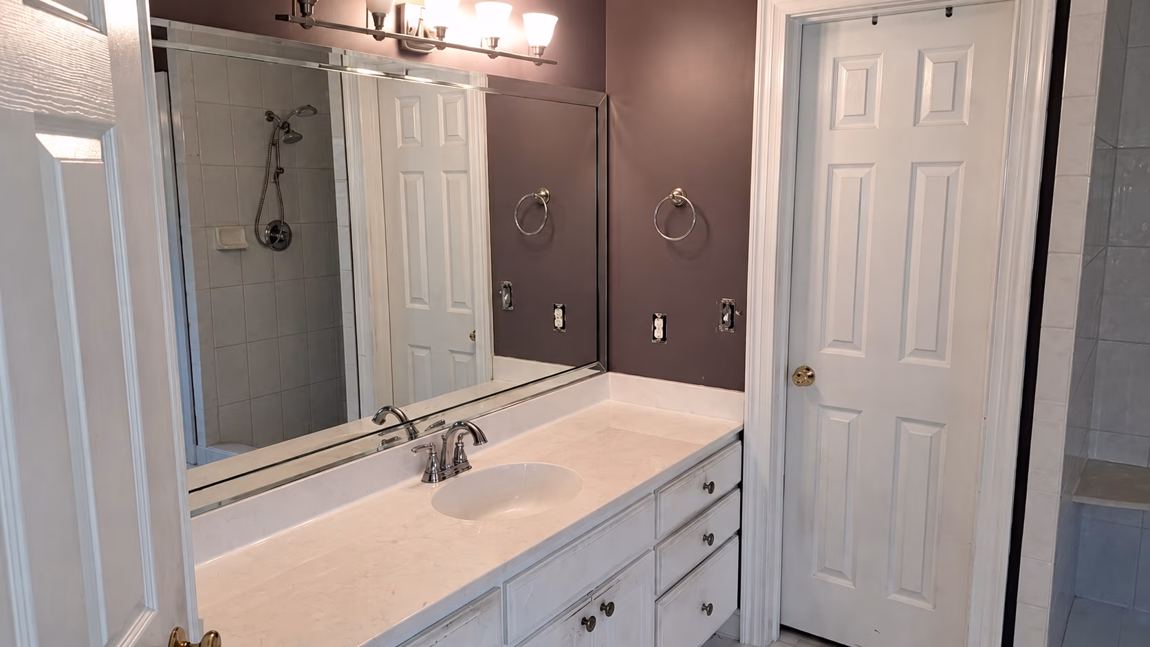 Bathroom vanity with white countertop, sink, large mirror, purple walls, and white door beside a tile shower.