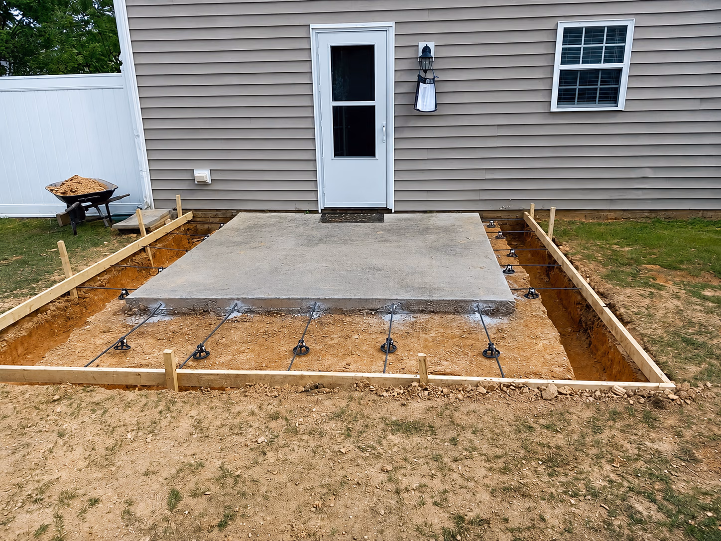 Concrete slab patio under construction with rebar and wooden frame next to house door and window.