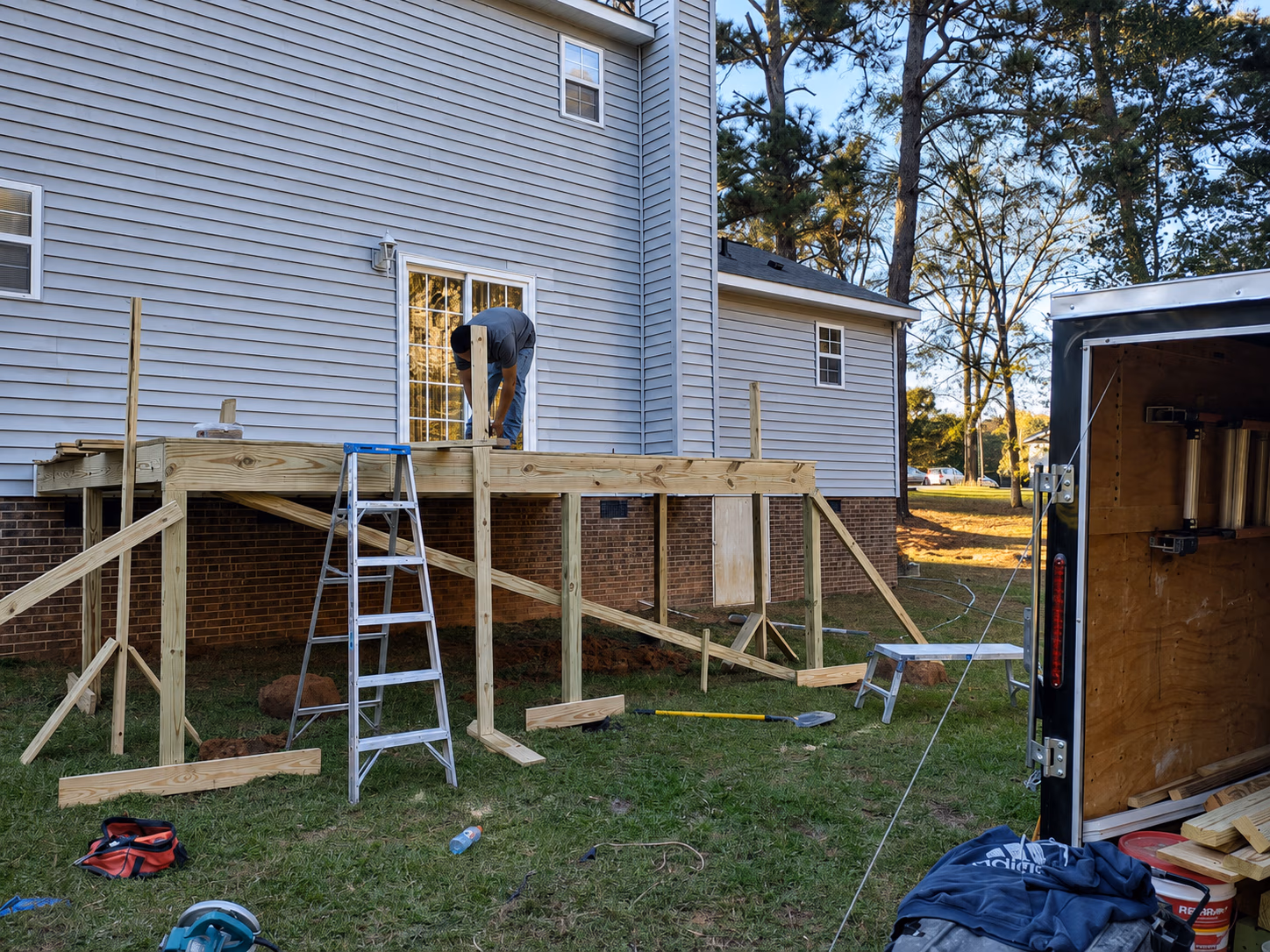 Man working on wooden deck frame attached to house with tools and trailer in a grassy backyard.