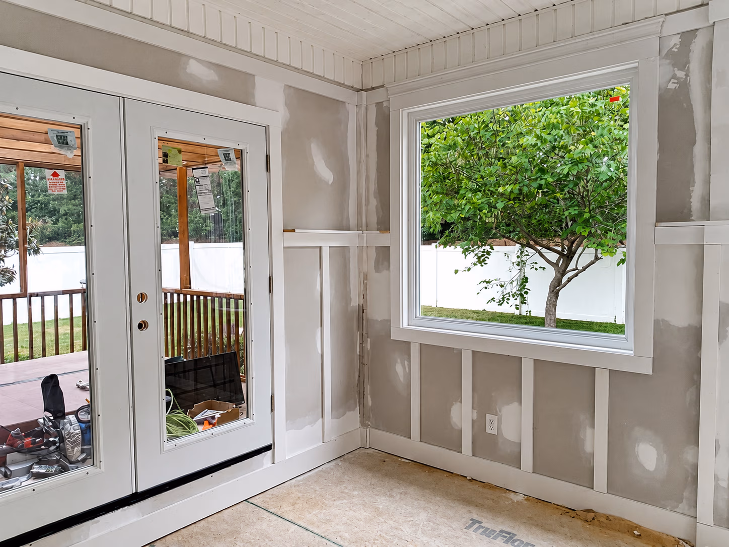 Interior room with unfinished drywall, large window showing green tree, and glass-paneled double doors.