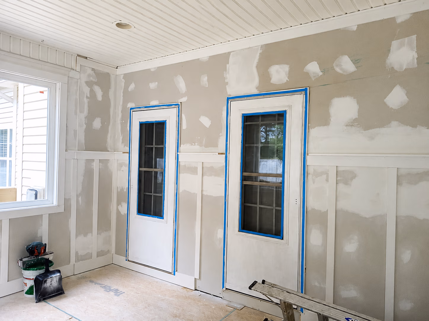 Interior room under renovation with drywall, two taped door frames, window, and tools on floor.