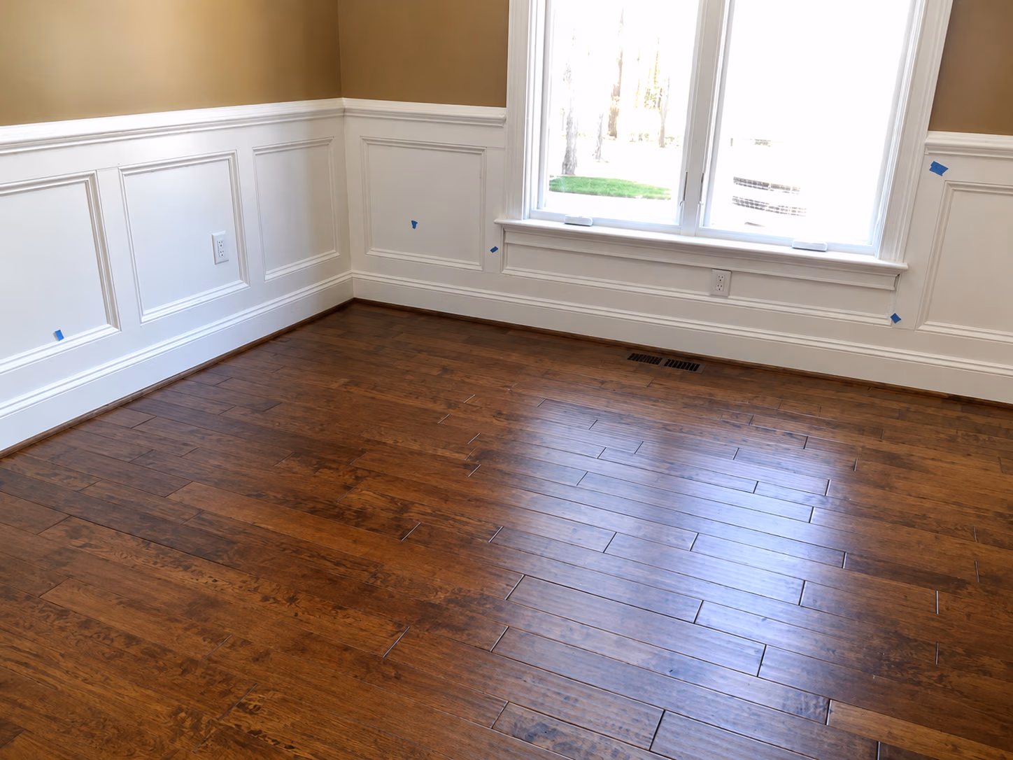 Empty room corner with white wainscoting, tan walls, brown hardwood floor, and bright window light.