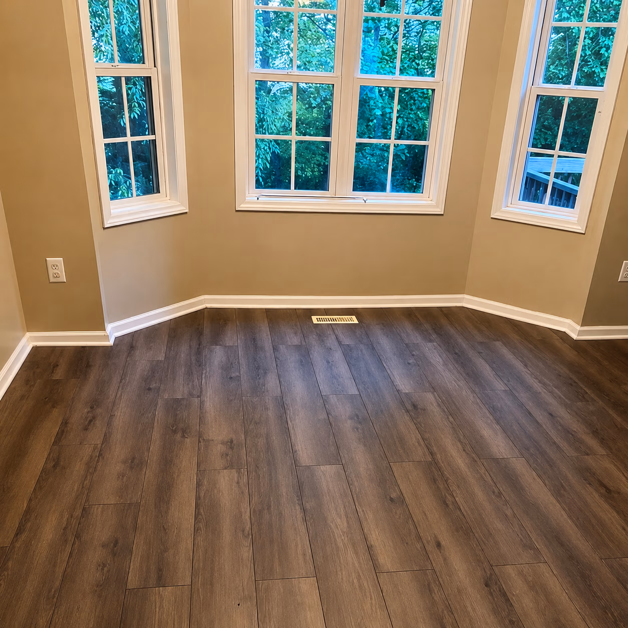 Empty corner room with wood floor, beige walls, white trim, and three windows showing green trees outside.