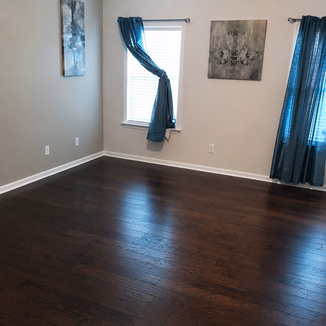 Empty room with dark hardwood floor, neutral walls, two windows with blue curtains, and two wall paintings.