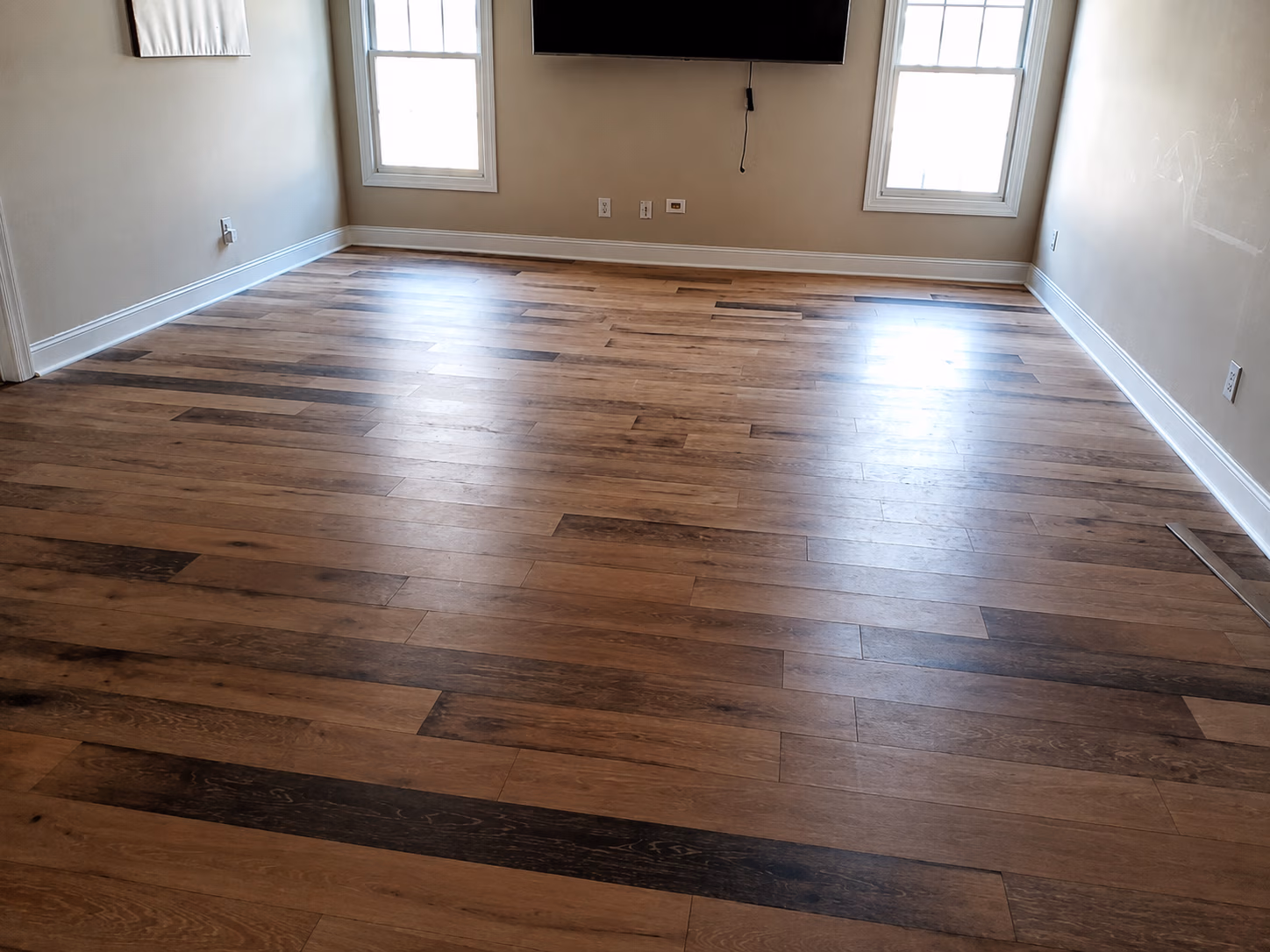 Empty room with light wood flooring, beige walls, two windows, and a wall-mounted TV.