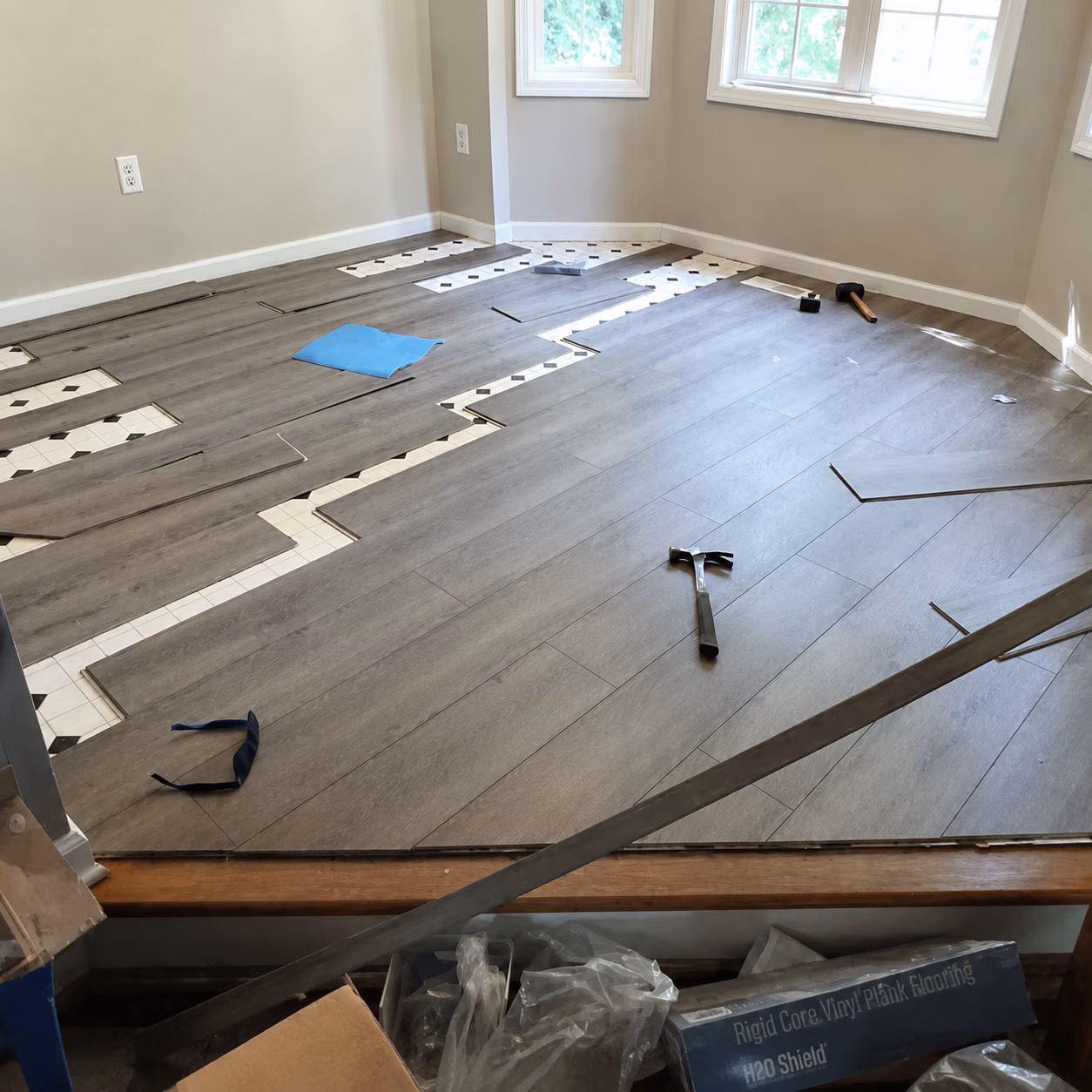 Partial installation of gray wood plank vinyl flooring over old tile with tools scattered around.