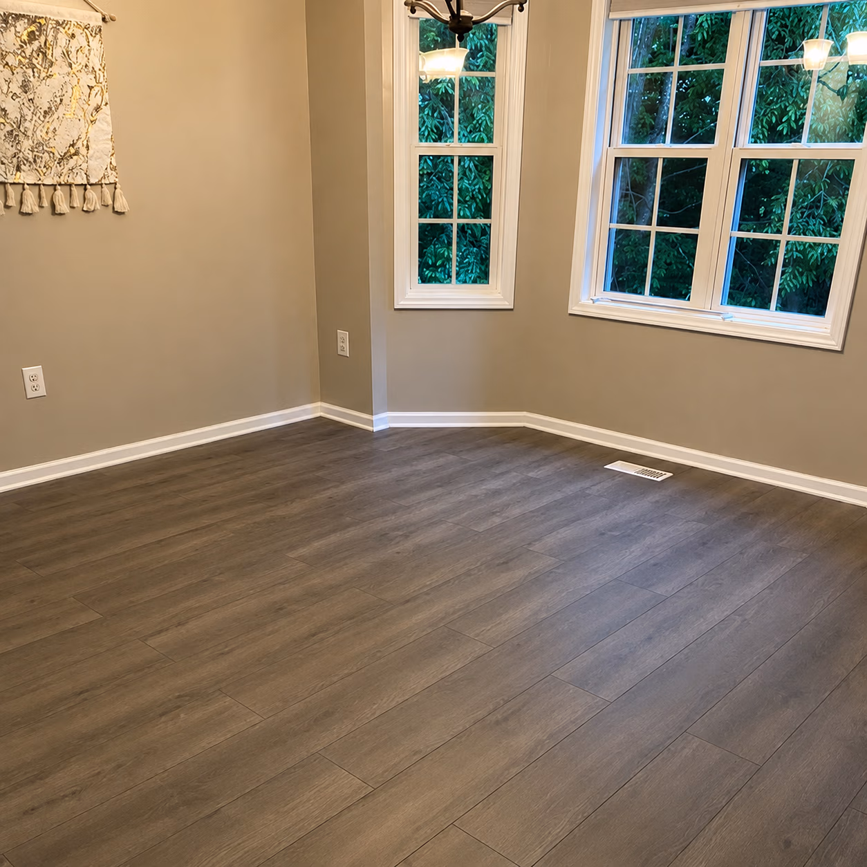 Empty room with gray wood laminate flooring, beige walls, three windows and white baseboards.