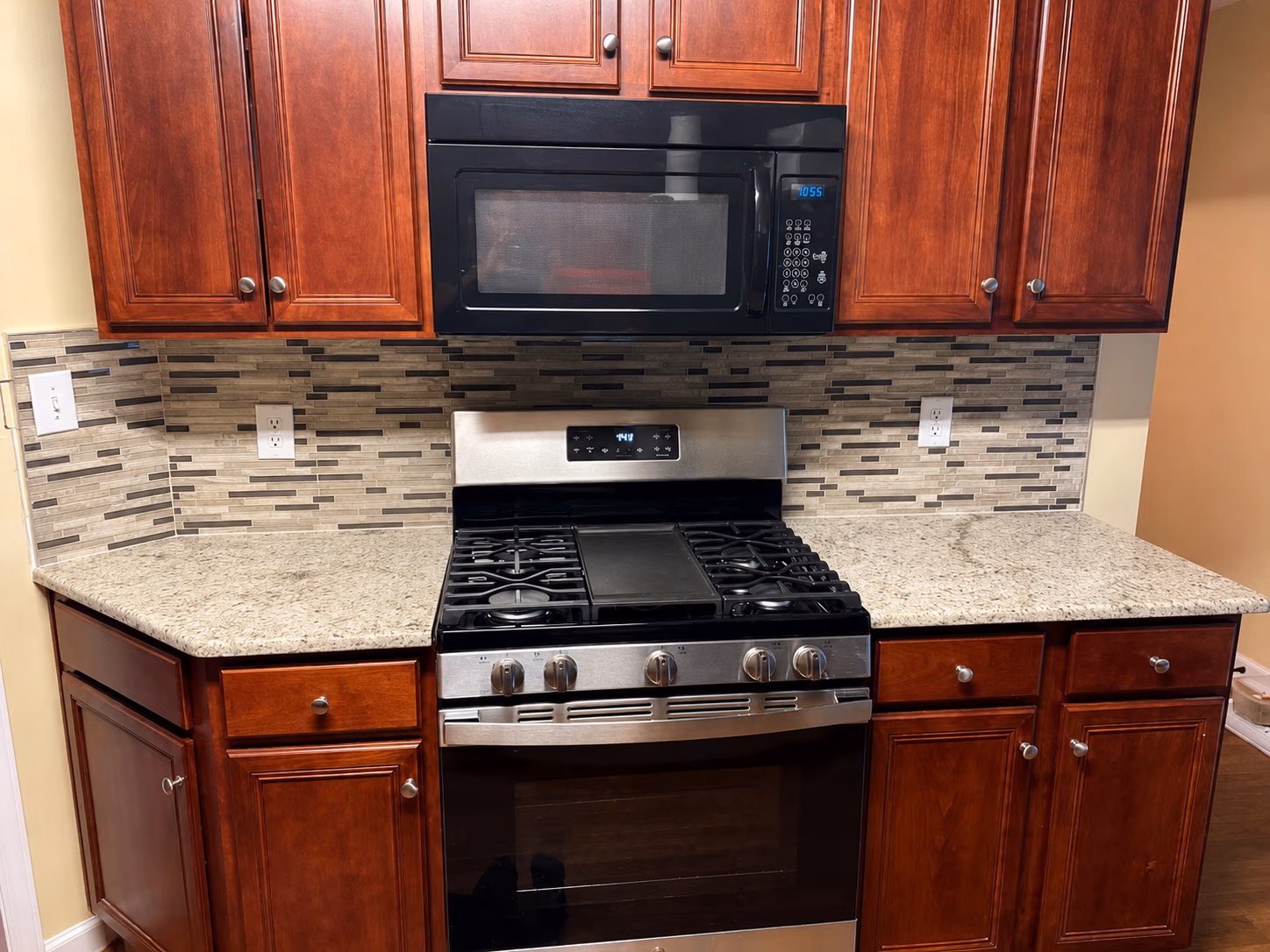 Kitchen stove with gas burners, stainless oven, black microwave, brown cabinets, and granite countertops.