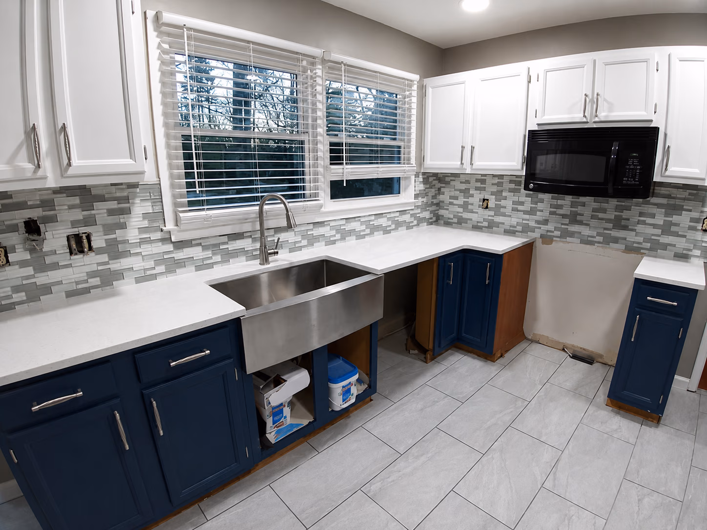 Kitchen with blue and white cabinets, large stainless steel sink under windows, and gray tile floor.