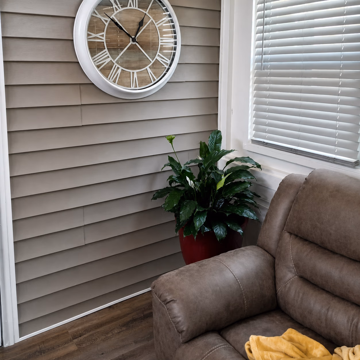 Brown leather recliner near window blinds, large wall clock, and a green potted plant in red pot.