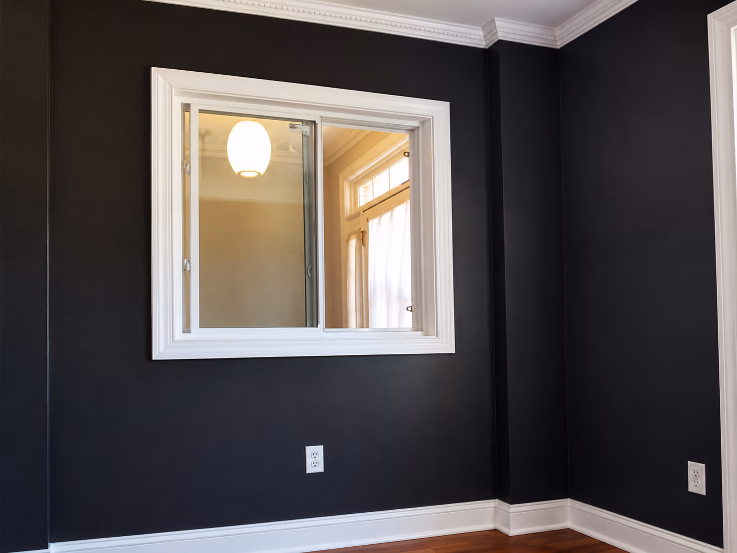 Empty room with dark walls, white trim, hardwood floor, a window showing a lit hanging light inside.