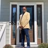 Man wearing beige blazer and sunglasses standing in front of a house door with glass panels.