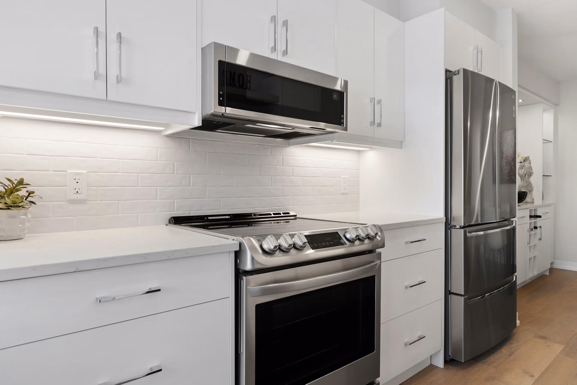 Modern kitchen with stainless steel stove, microwave, refrigerator, white cabinets, and wood floor.
