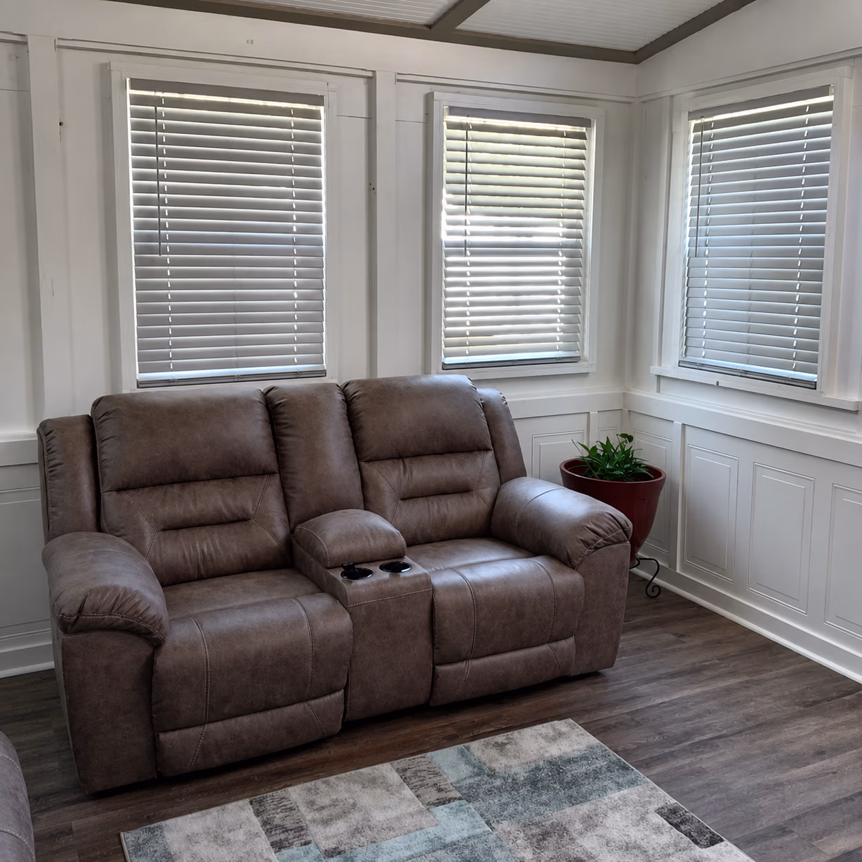 Brown leather reclining loveseat with cup holders in a corner room with white walls and three windows.