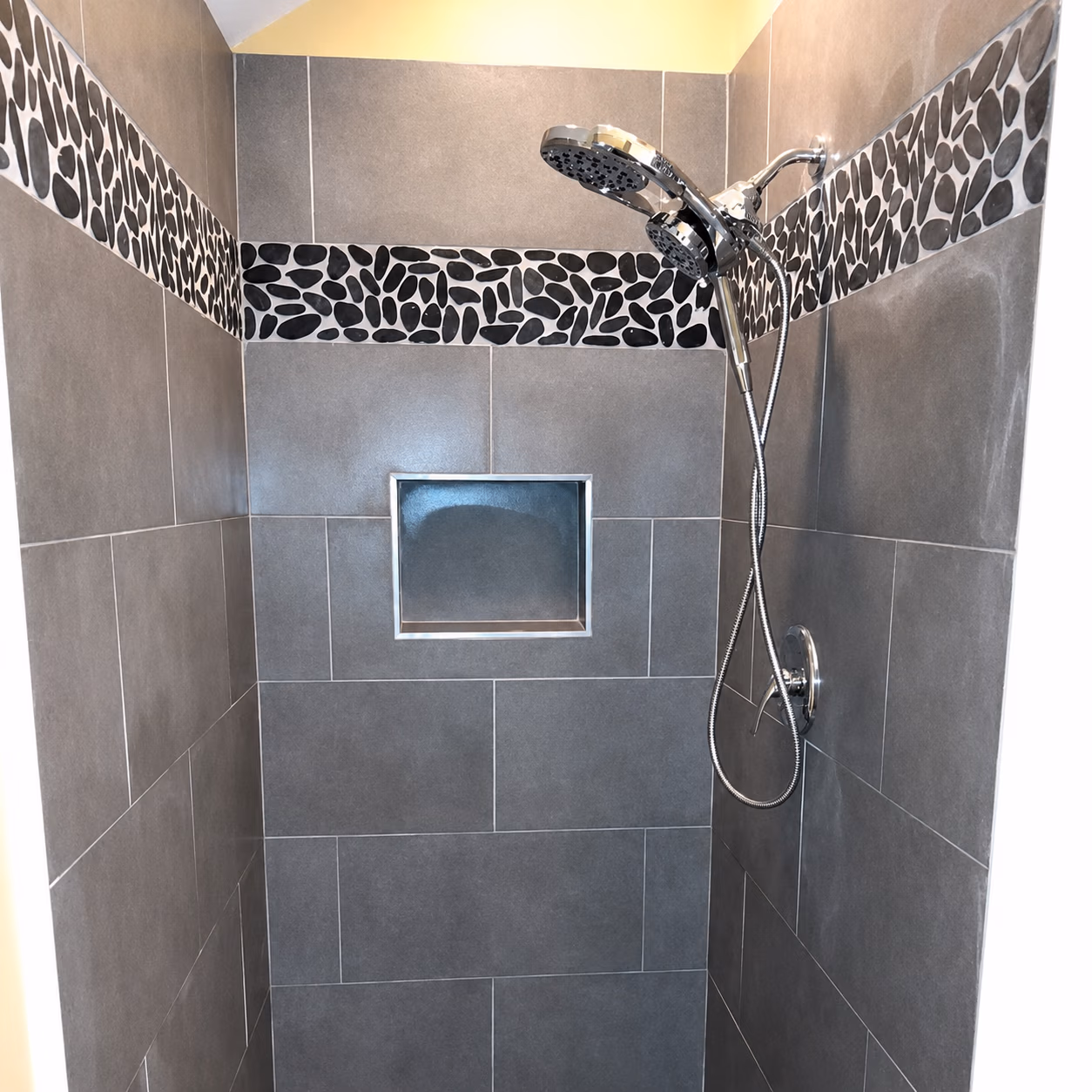 Modern shower with gray tiles and a decorative black pebble border near chrome showerhead.