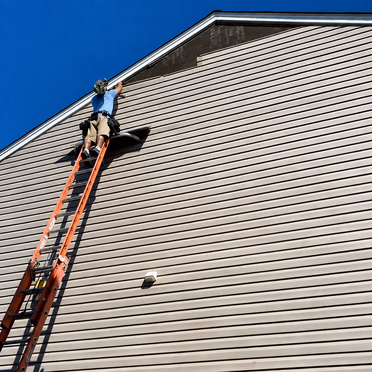 Worker on ladder installing or repairing siding near the roof peak of a house under clear blue sky.