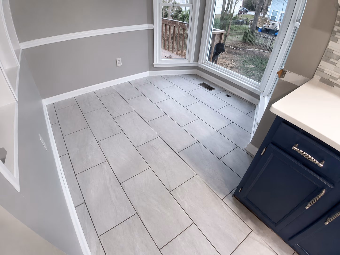 Empty room with gray tiled floor, gray walls, large corner window, and blue kitchen cabinets.