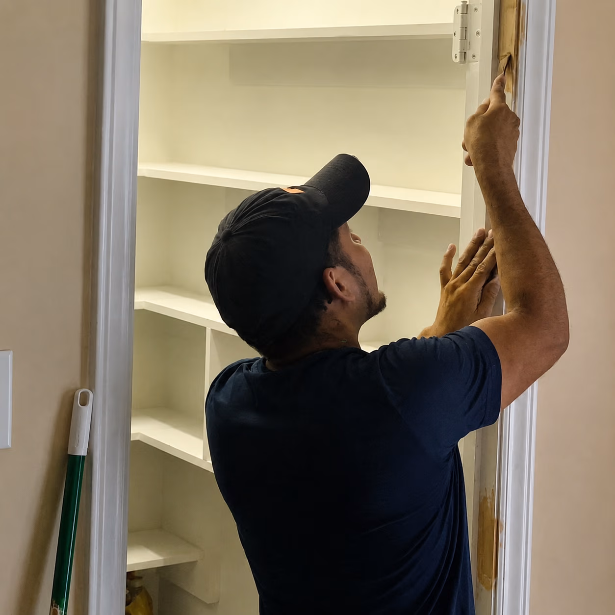 Man in black cap applying wood stain or varnish to a white door frame.